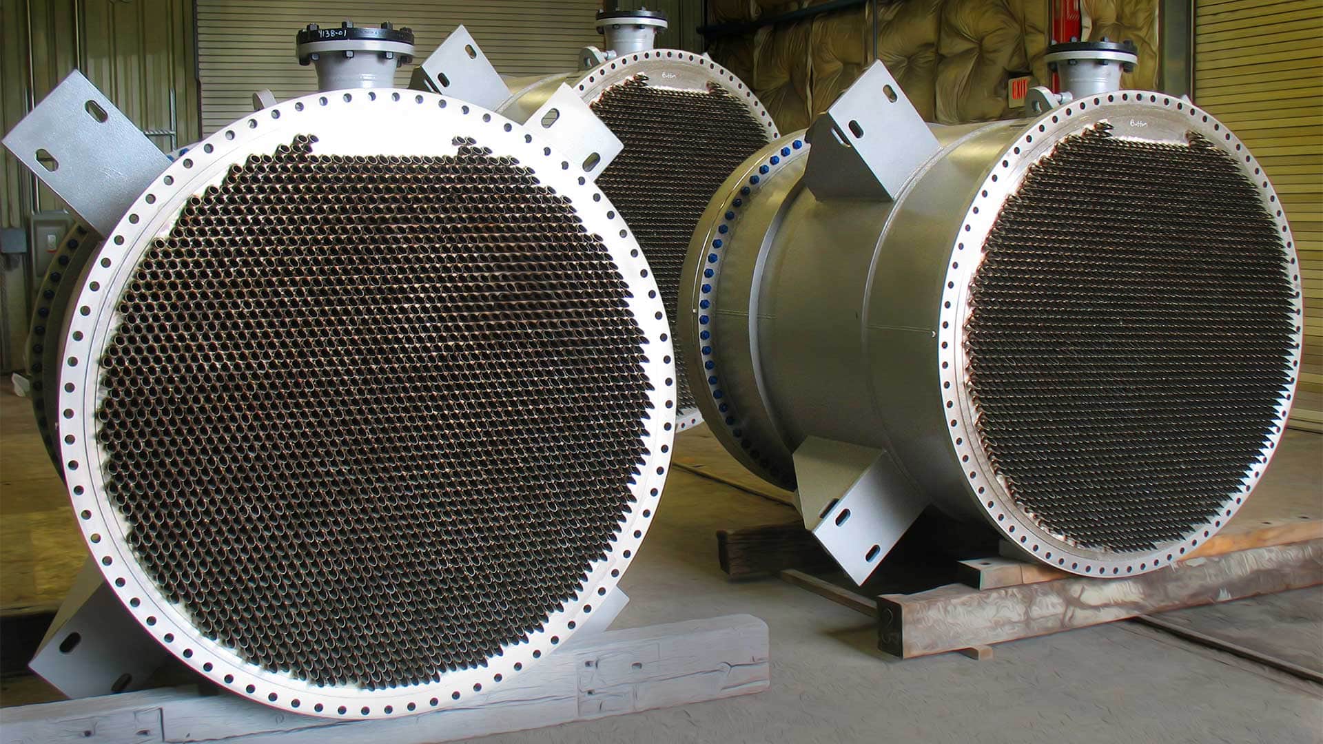 Row of large shell-and-tube heat exchangers with dense circular tube bundles visible through their front flanges, staged in a fabrication shop for a PVC reactor condenser order.