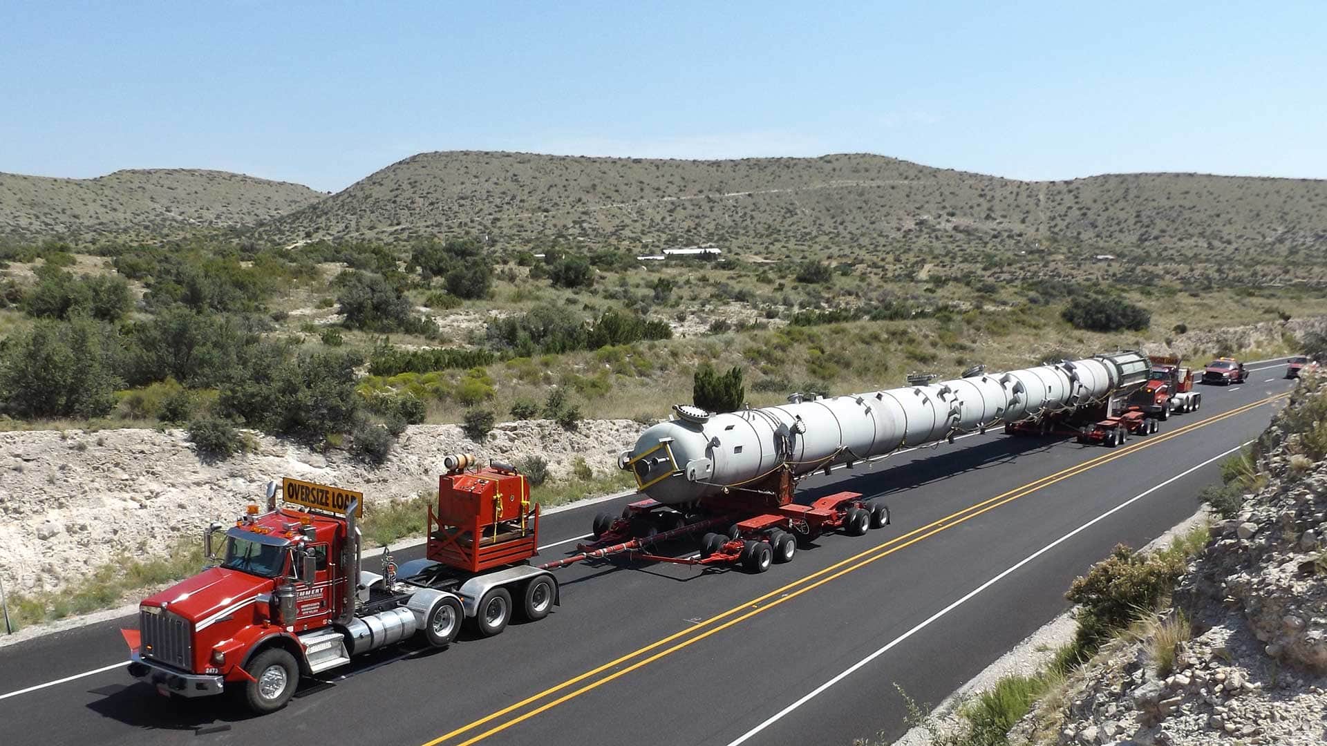 Oversize load truck hauling a large white cylindrical vessel down a two-lane highway through a desert landscape, escorted by a lead truck marked "Oversize Load" and trailing vehicles.