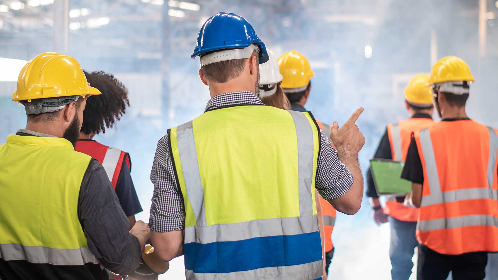 Group of construction workers wearing safety vests and hard hats walking through an industrial facility.