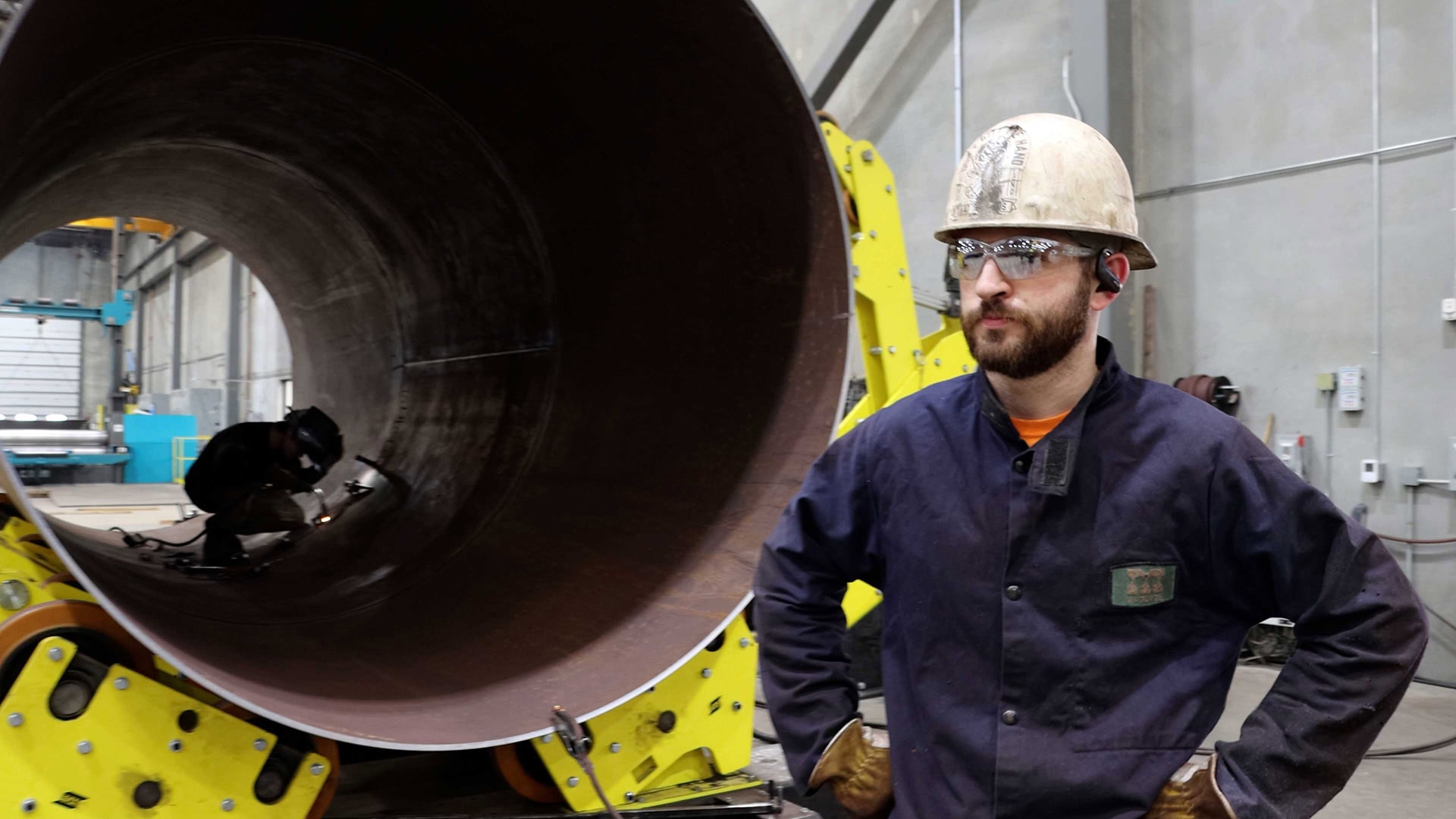 Worker in hard hat and safety glasses standing near a large steel pipe as another worker welds inside.