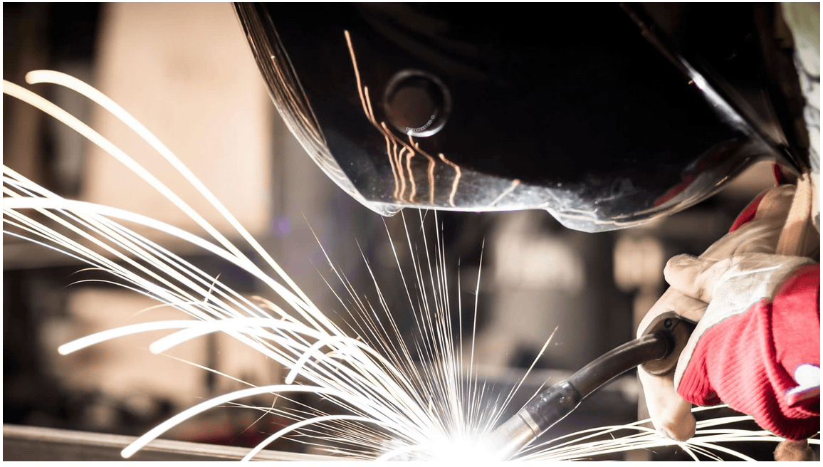 Close-up of a welder performing industrial welding in a steel fabrication shop, wearing protective gloves and helmet as bright sparks fly from the metal.