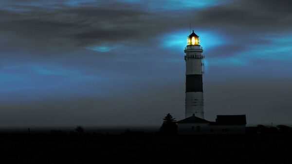 Lighthouse glowing at dusk with its beacon shining through dark clouds over the ocean.