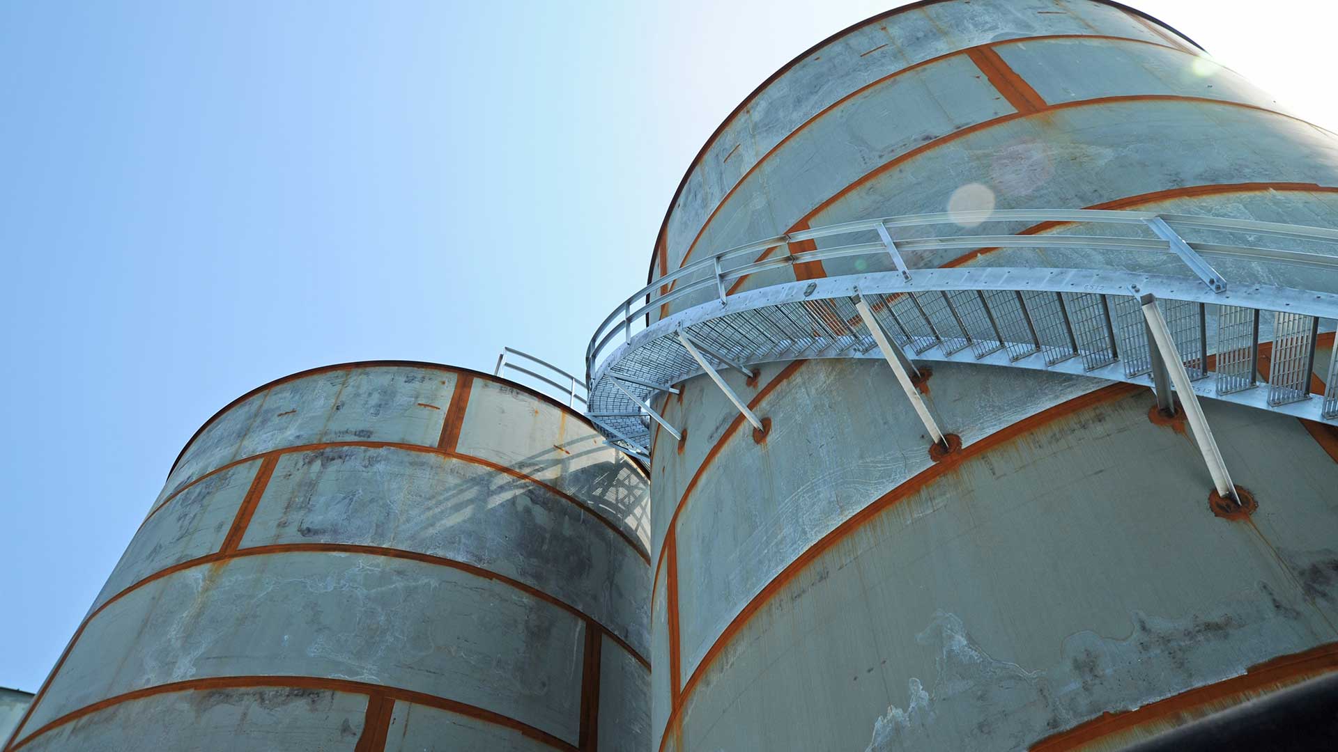 Two large steel storage tanks with visible weld seams and an exterior access walkway viewed from below.