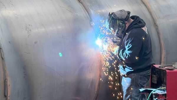 Welder wearing protective gear welding a large steel tank as sparks fly.