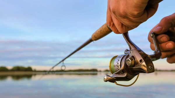 Hand holding a fishing rod and reel extended over calm water at sunrise.