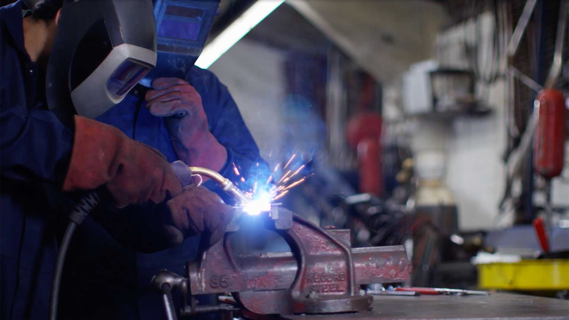 Welding student performing MIG welding on a metal component secured in a bench vise inside a fabrication shop.