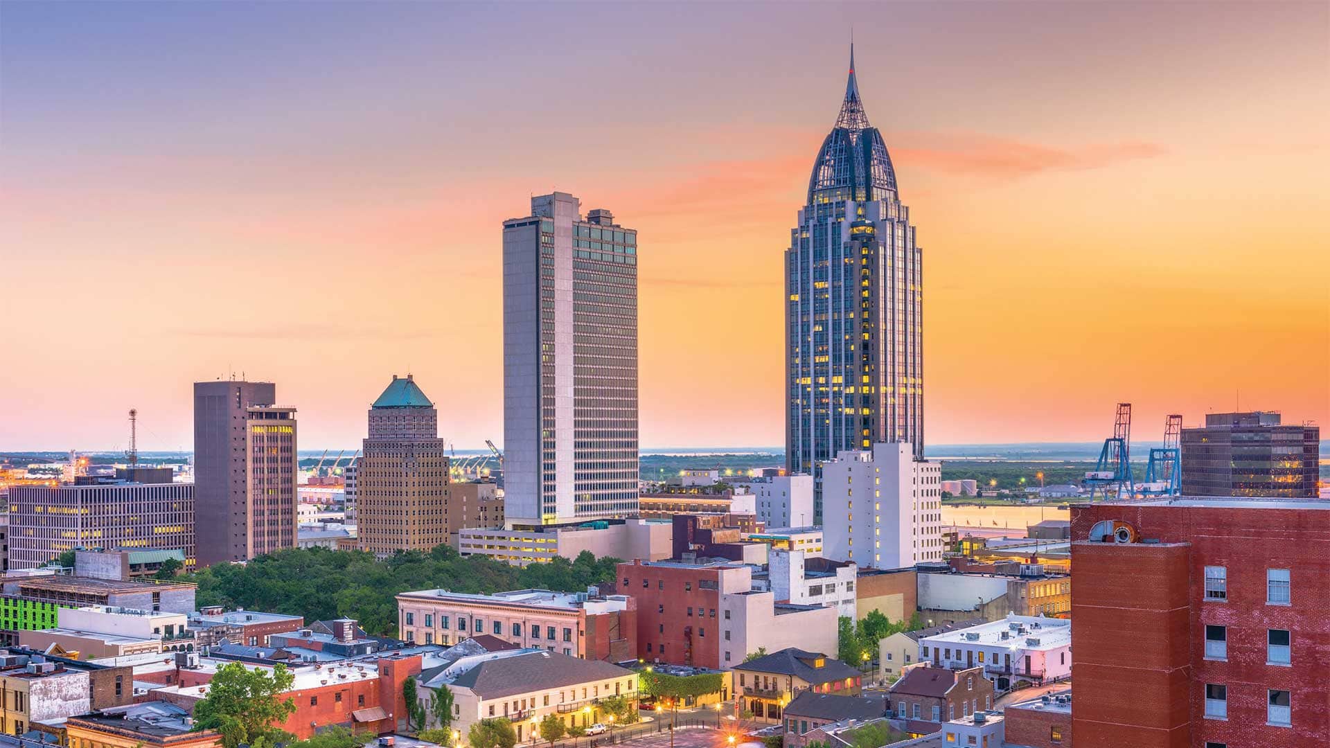Downtown Mobile, Alabama skyline at sunset featuring high-rise buildings and the waterfront in the background.