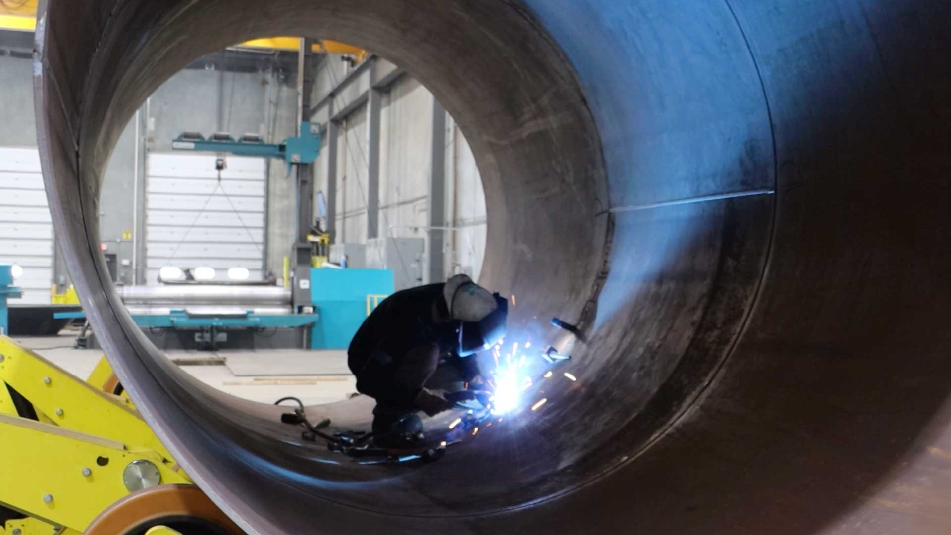Welder working inside a large rolled steel shell in a fabrication facility.