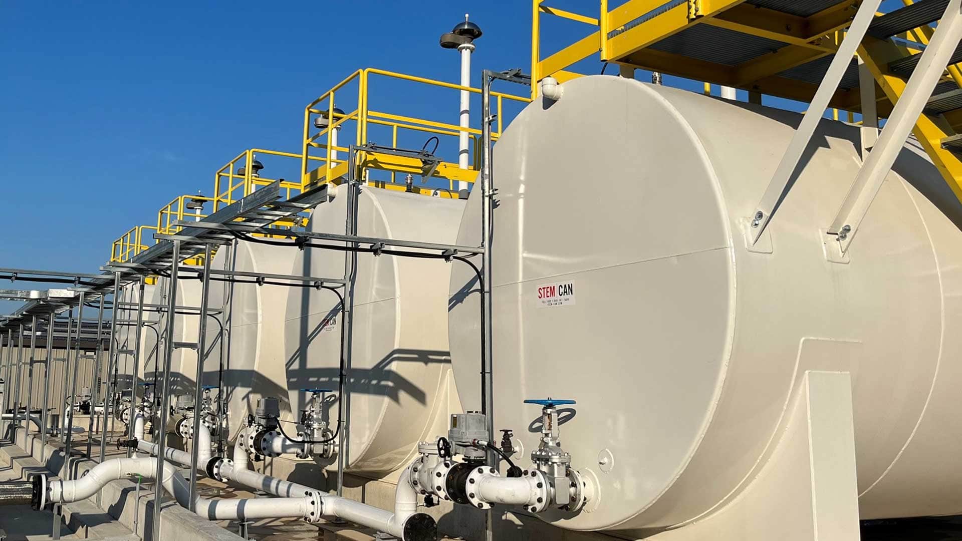 Row of STEM-CAN steel fuel storage tanks with interconnected piping, valves, and elevated access platforms at an industrial facility.