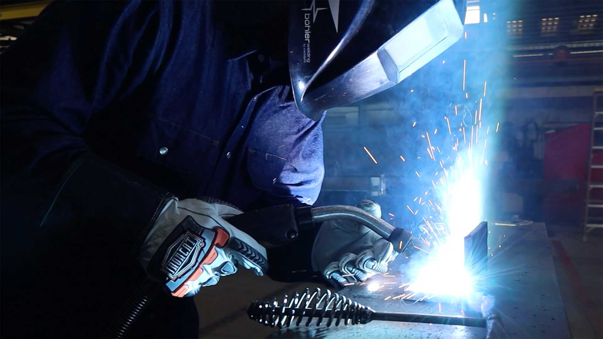 Welder performing MIG welding on a steel plate with bright arc and sparks in a fabrication shop.