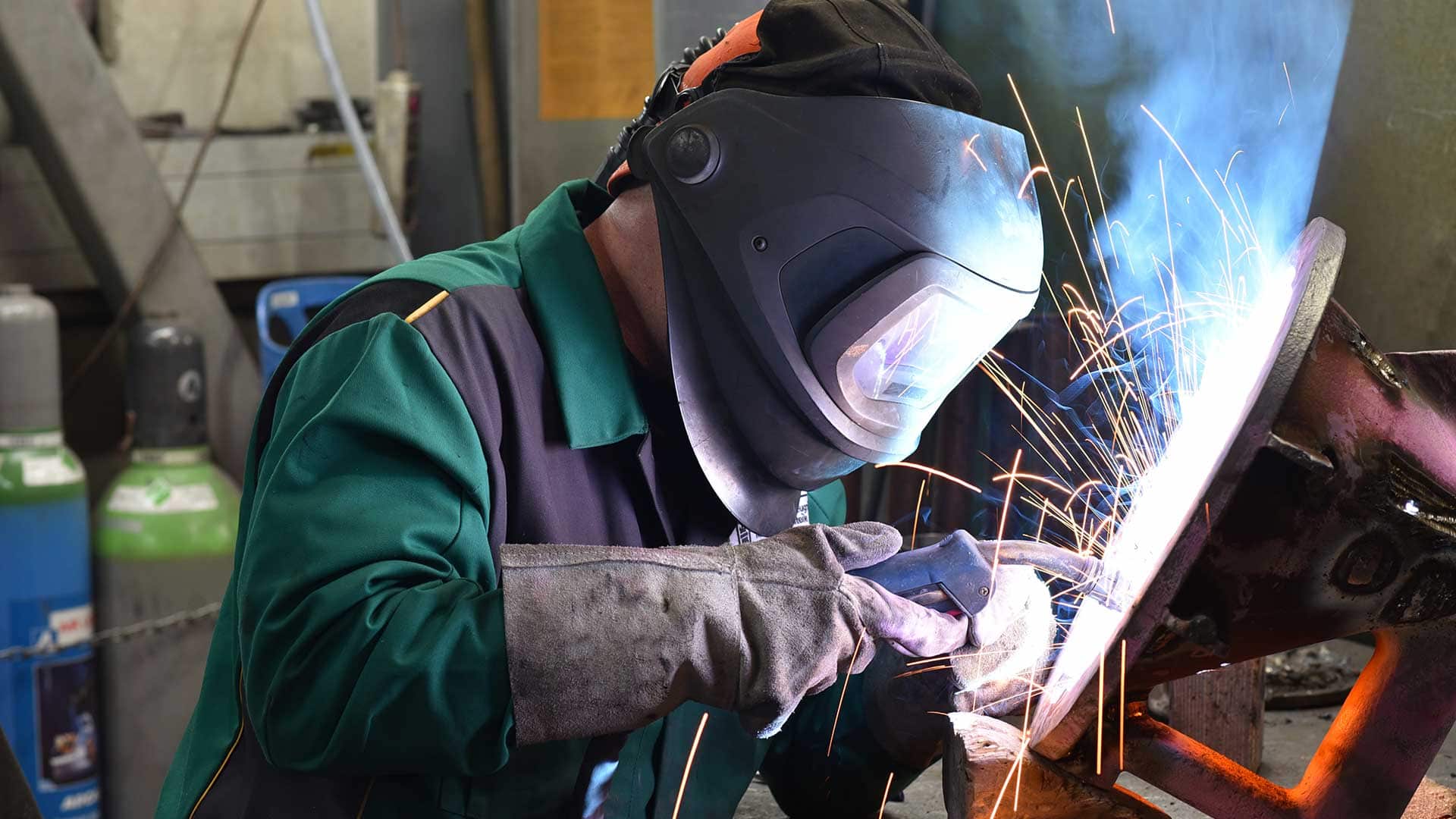 Welder performing arc welding on a steel component with sparks in a fabrication shop.