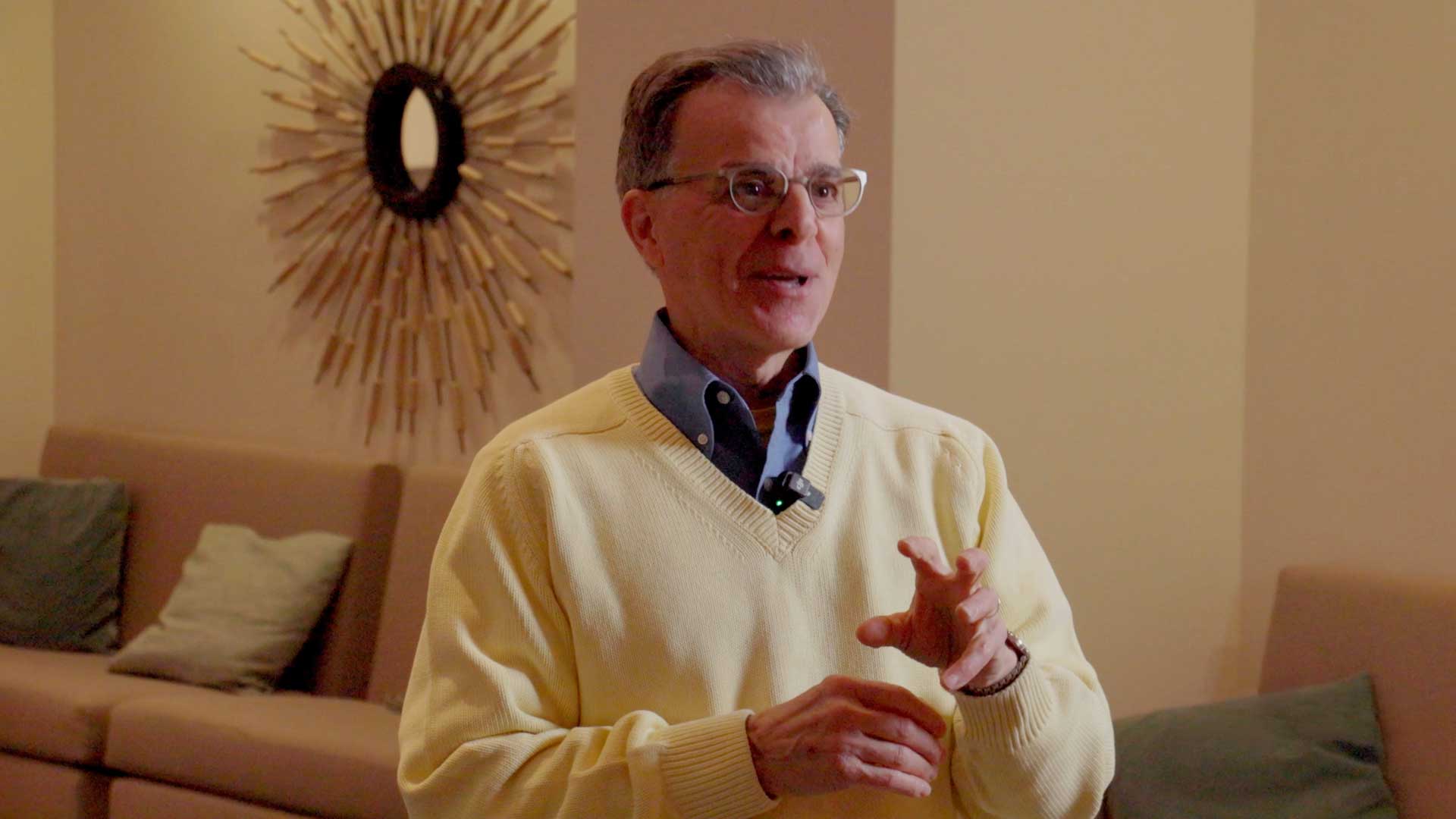 Man speaking during an interview, wearing glasses and a light sweater, seated indoors.