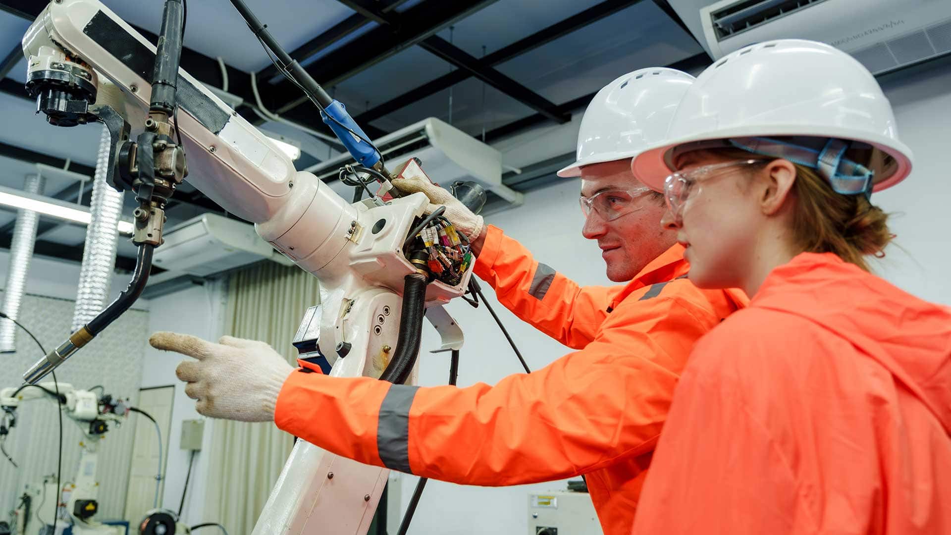 Two workers wearing hard hats, safety glasses, and high-visibility clothing inspect and operate a robotic welding arm in an advanced manufacturing facility.