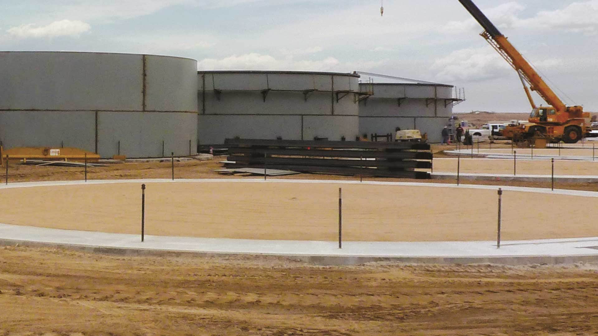 Field-erected steel storage tank foundations under construction with nearby tanks, crane equipment, and construction materials.