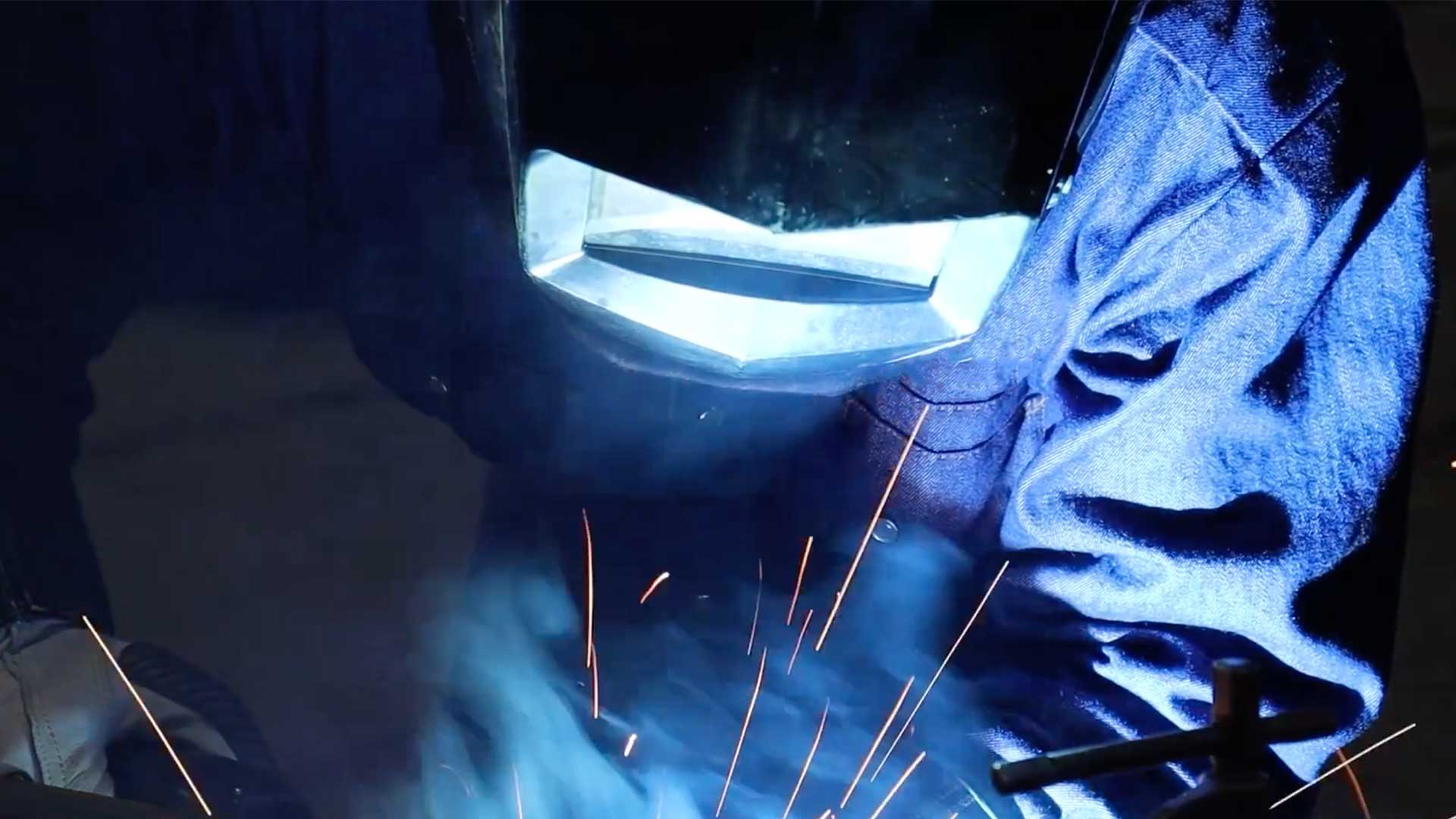 Close-up of a welder wearing protective gear while performing steel fabrication work with sparks and arc light visible.