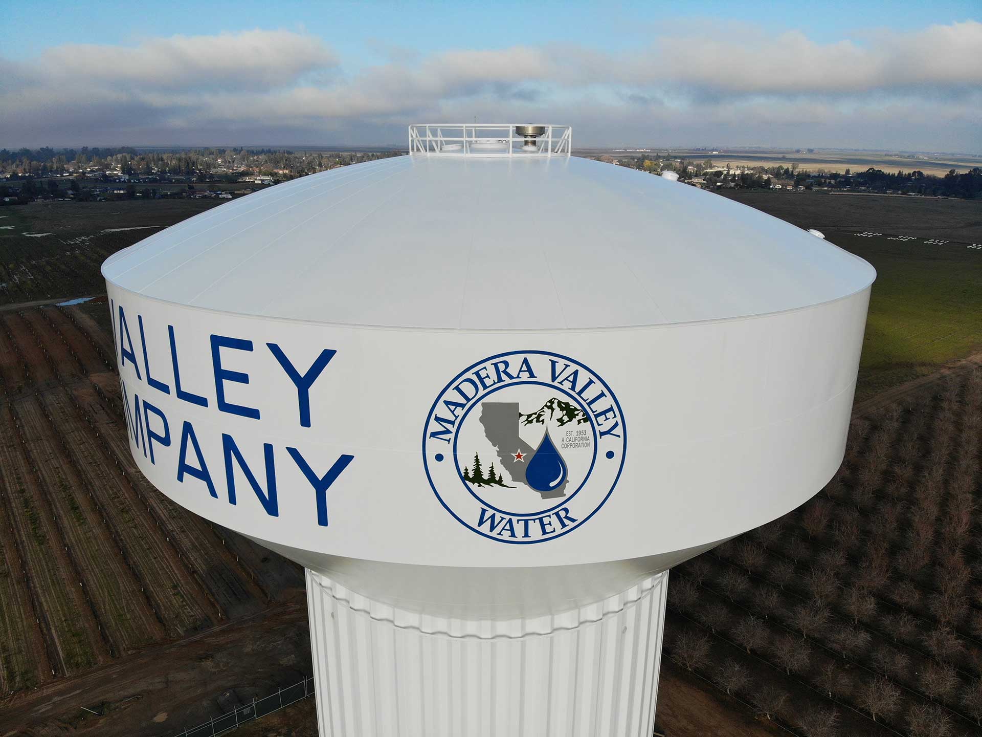 Aerial close-up of the white 1.5-million-gallon Madera Valley Water elevated water tank, showing its logo and lettering, overlooking surrounding farmland.