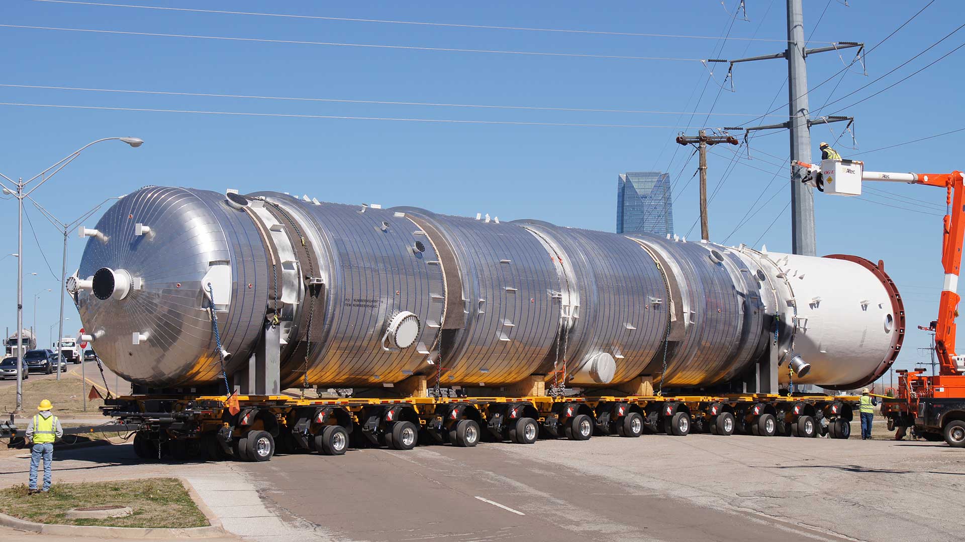 Large insulated stainless steel diesel recovery tower being transported on a multi-axle self-propelled modular trailer through a city street, with a utility bucket truck lifting power lines to clear the load.