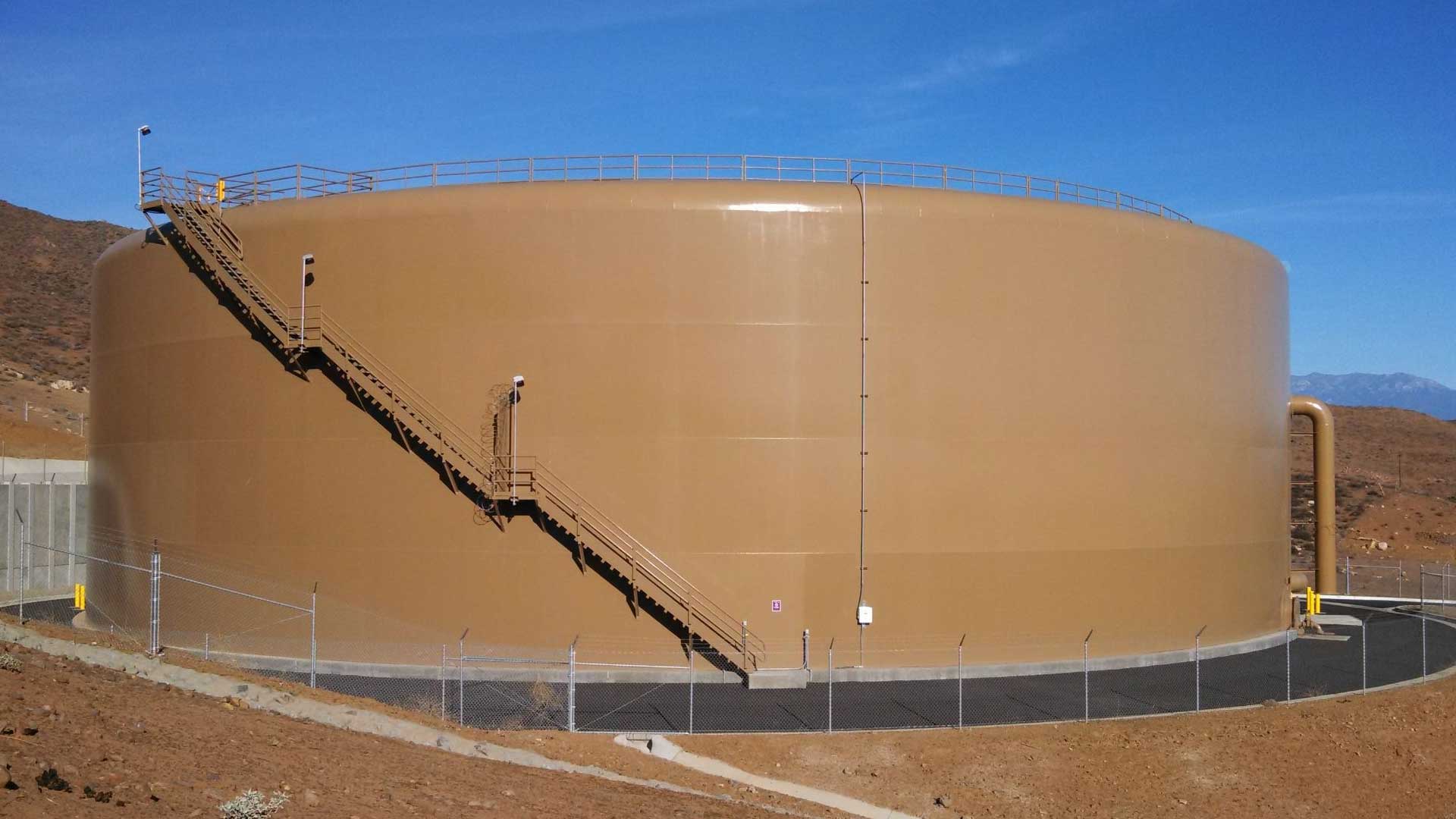 Large brown steel water storage tank with a diagonal exterior staircase, surrounded by chain-link fencing, set against a desert hillside and blue sky near Winchester, California.