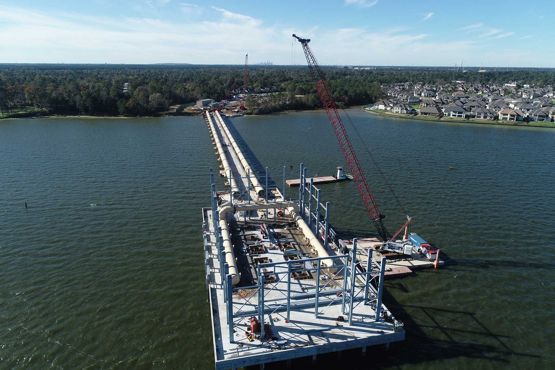 Aerial view of an intake pump station under construction on a large reservoir, with large intake pipes on trestle supports extending across the water, a barge-mounted crane, and a residential shoreline in the background.