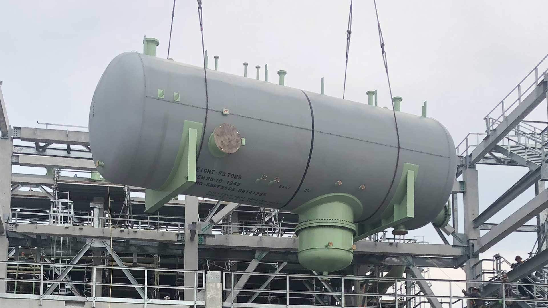 Gray horizontal pressure vessel weighing 53 tons being lifted by crane cables into a multi-level steel structural framework at an industrial construction site.