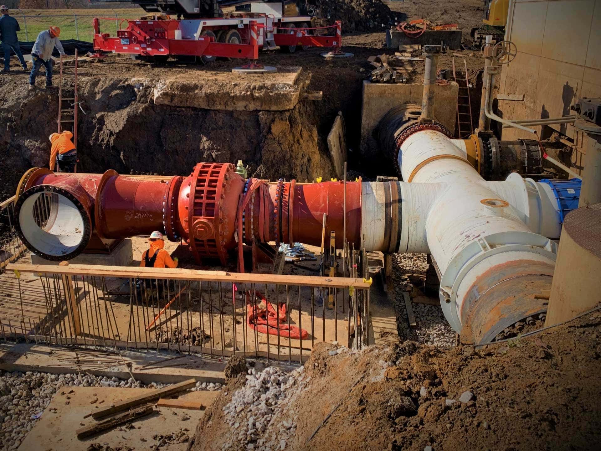 Workers in an excavated trench connecting a large red steel pipe assembly with flanged fittings to an existing white water main as part of the Bird Creek Pump Station tie-in repair project near Tulsa.